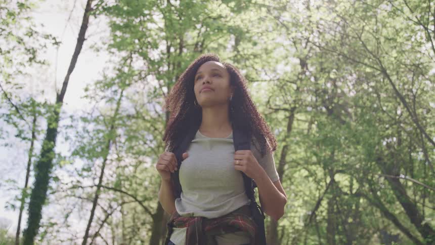 Black woman with curly hair and a backpack walks and looks at tall trees in the forest