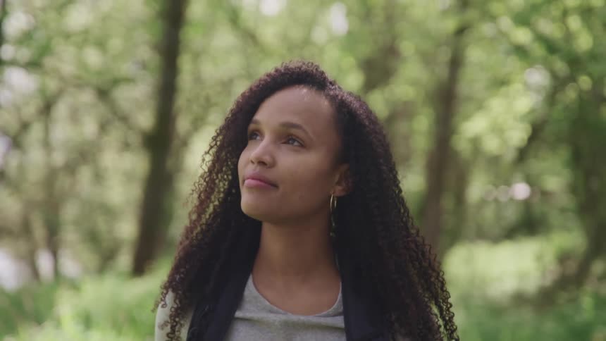 Black woman with curly hair a grey t shirt and earrings walks and looks at tall trees in the forest