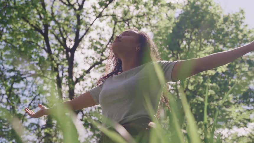 Woman with arms wide open looks up with eyes closed while standing on the grass near the trees