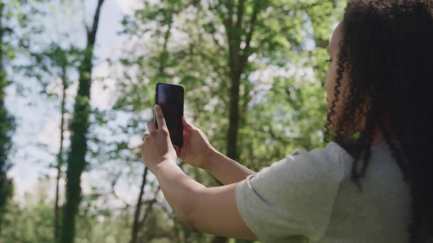 Woman with curly hair and a grey t shirt takes a panorama photo with a smartphone in the forest