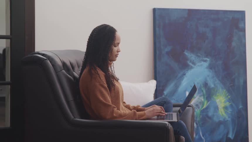 Black woman with curly hair and a brown sweater sits in a black armchair next to an oil painting and uses a laptop in a room