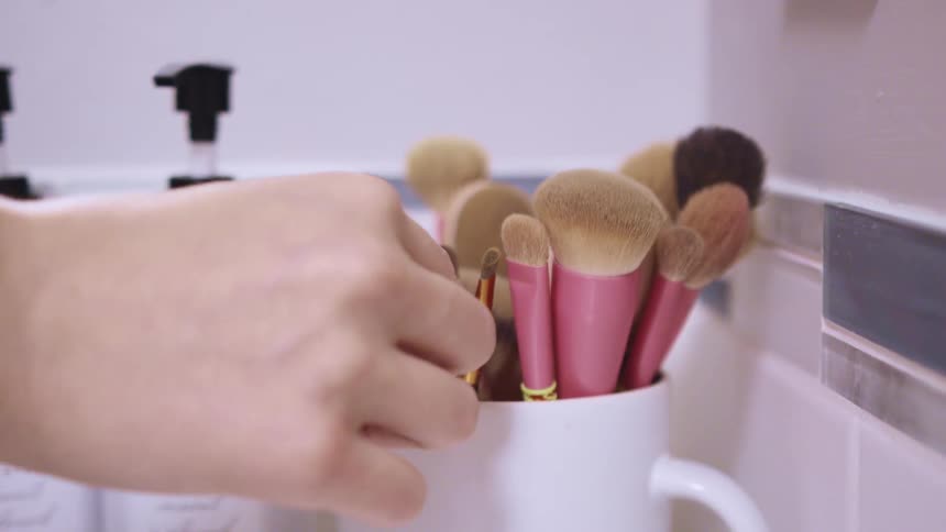 A woman grabs a makeup brush from a mug full of makeup brushes on her bathroom counter