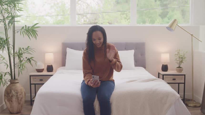 Female sits on the end of a bed as she smiles and waves while video chatting using her smart phone