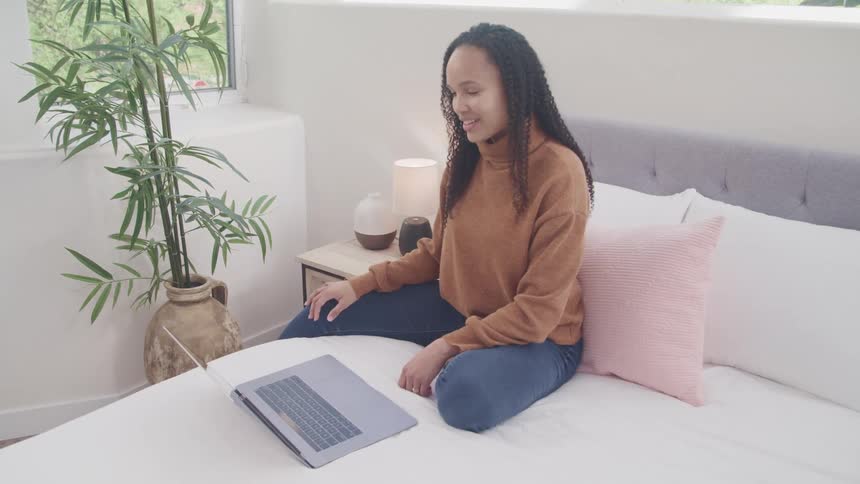 Black woman sits on a bed and looks at a laptop smiling and waving