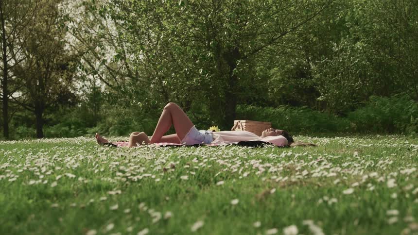 Woman laying over a blanket in the grass near a picnic basket surrounded by white wild flowers