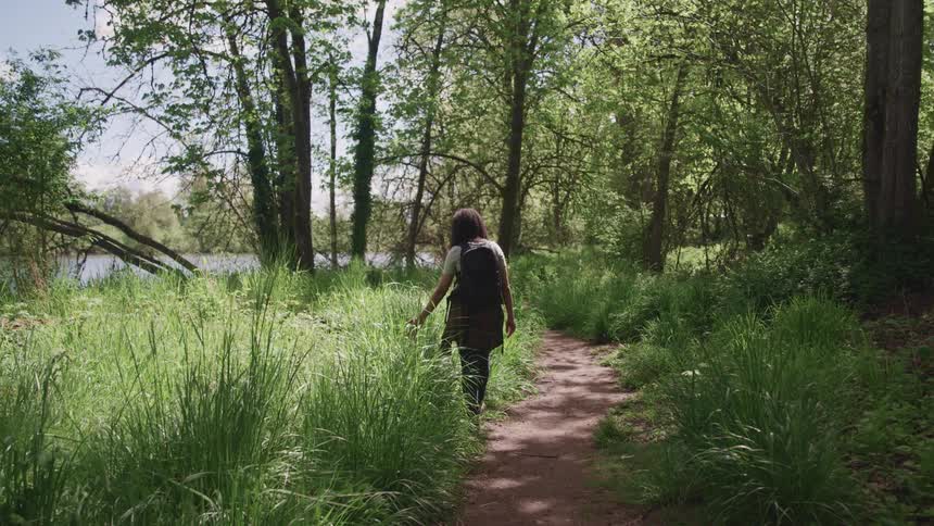 A woman runs her hand through the tall grass as she walks along a dirt path through a woodland
