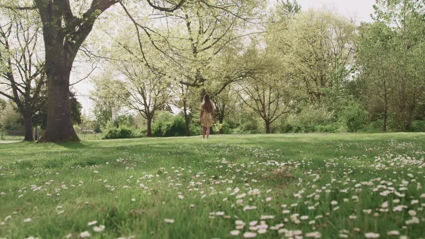 Woman with a yellow dress and brown hair holds a bunch of sunflowers and walks on green grass in the park