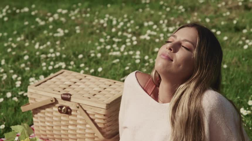 A woman sits facing the sky with her eyes closed in a meadow next to a picnic basket