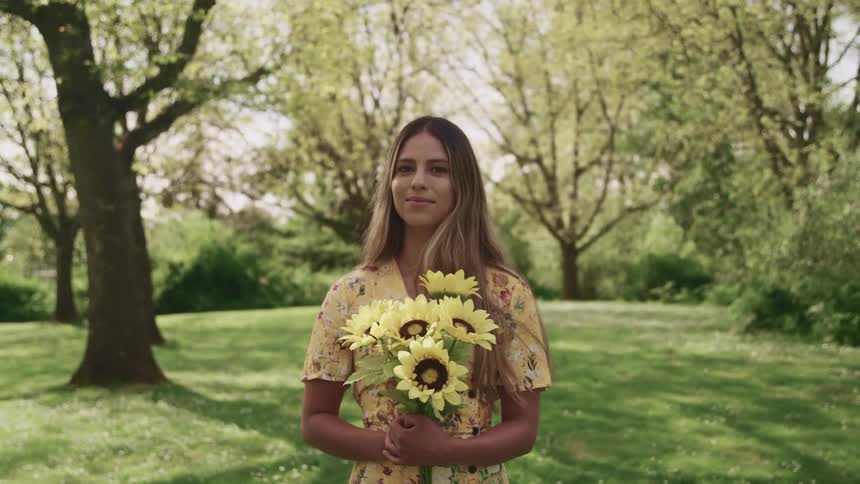 Young woman in a dress holds sunflowers in her hands standing on the lawn near the trees