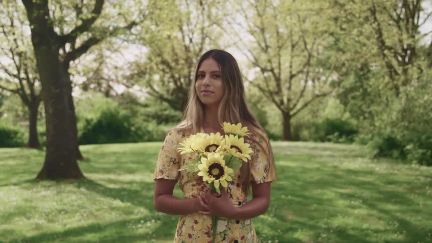Woman with brown hair a yellow dress and holding a bunch of sunflowers stands on grass and looks at the camera