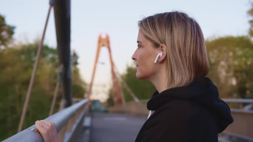 Woman wearing white earbuds stands on a bridge near a forest