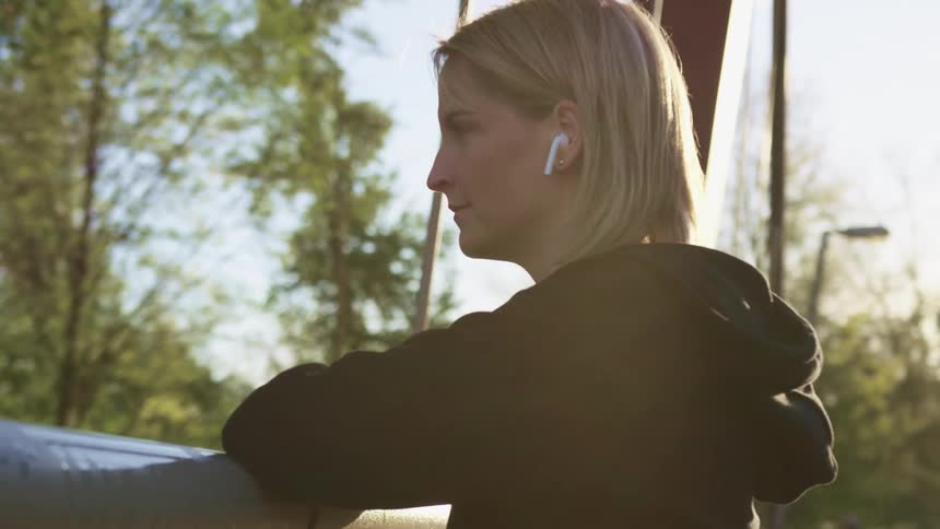A woman with ear buds in leans on a railing and looks off in to the distance