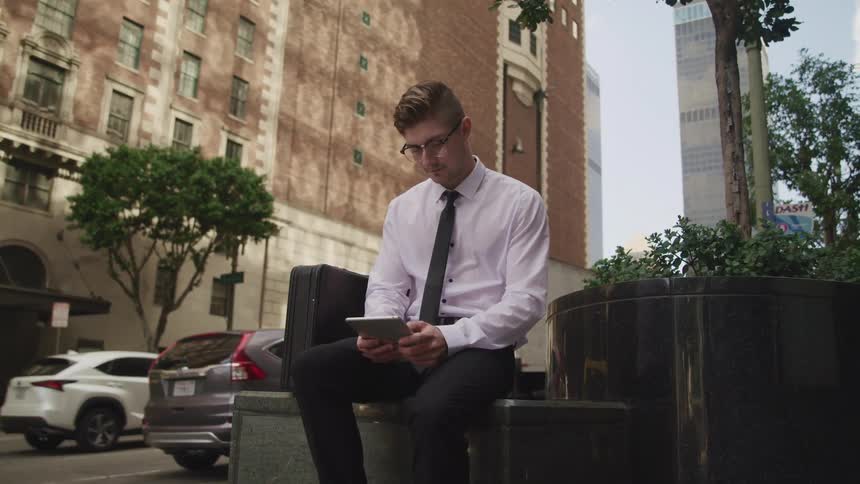 A man sits on a city street reading on his tablet as cars drive past