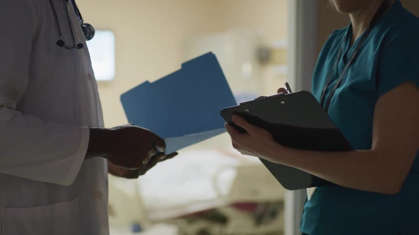 A nurse writes in a chart as she talks to a doctor outside of a hospital room
