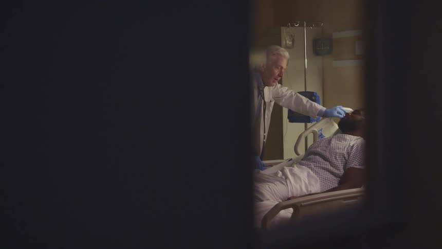 Through a small opening in a doorway an old doctor can be seen looking at a sleeping patient