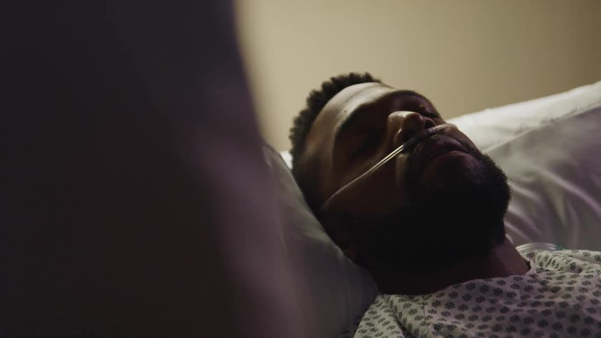 A patient sleeps in a hospital bed as a person stands by their bedside