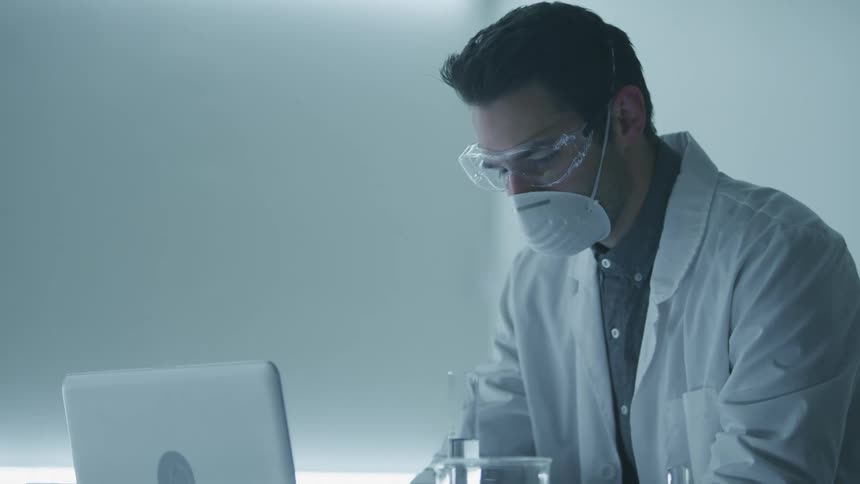 Scientist wearing lab coat and face mask works on a laptop in a laboratory