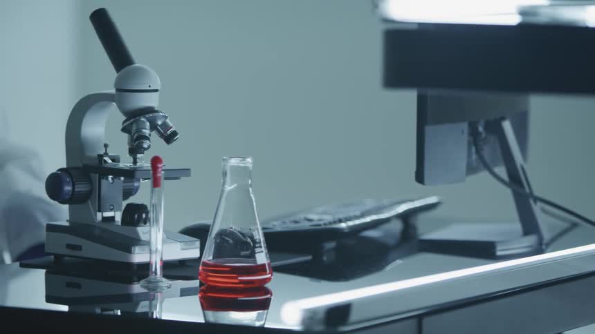 A microscope sitting on a computer desk next to a beaker of red liquid