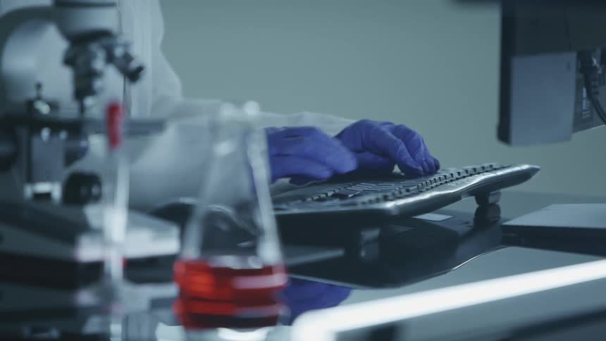 A scientist sits at a desk and types on a computer while wearing latex gloves