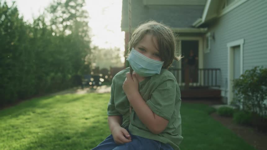 A boy wears a blue face mask as he poses on a rope swing in his yard as his mom watches from the back door