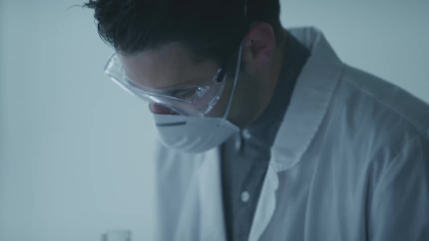 Doctor wearing goggles and face mask stands studying against pale colored walls