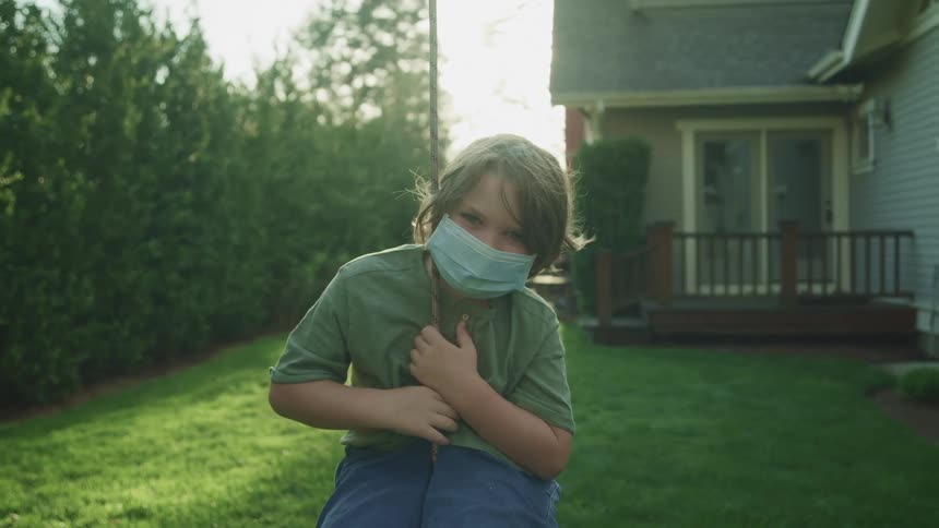 A boy in a medical mask looks into the camera while sitting on a swing in the backyard of a house during the day