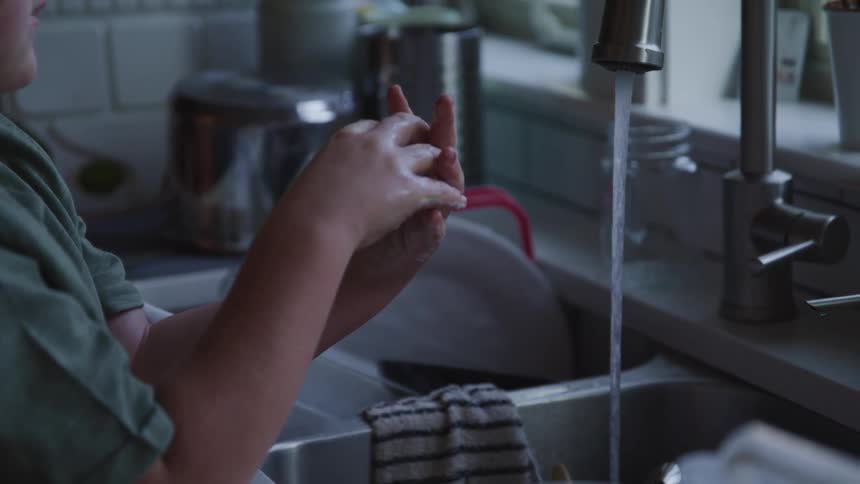 Boy with a green shirt puts his hands under a tap and washes his hands with water and soap in the kitchen