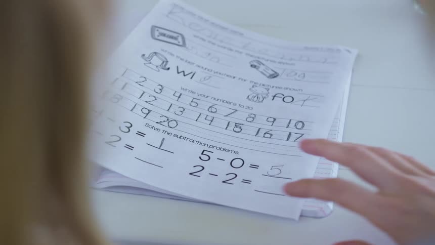 A girl uses a pencil to complete math problems on a worksheet as an adult checks her work