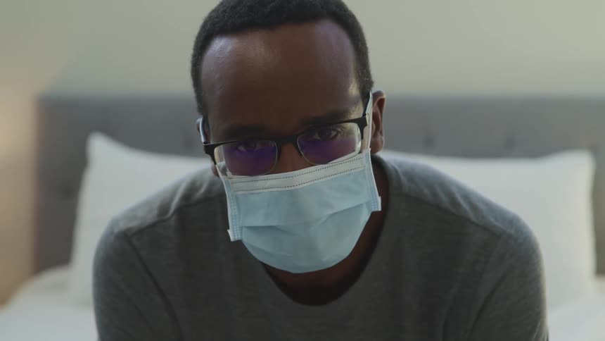 A man sits on a bed wearing a medical mask looking depressed