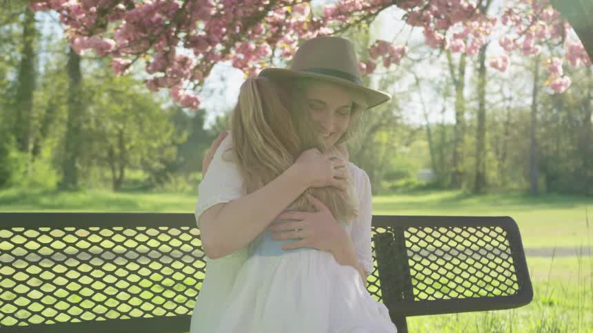 Mother sits on a park bench underneath a cherry blossom tree as she holds her daughter in her lap and smiles while giving her a hug