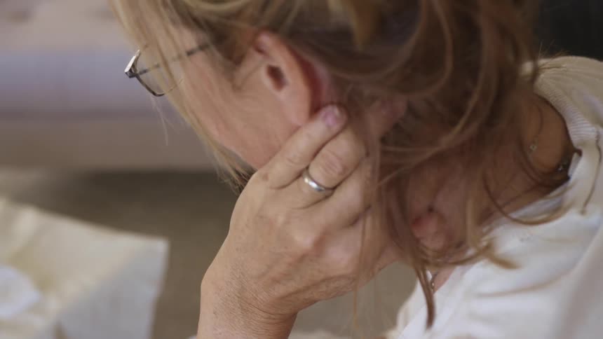 Middle aged woman with glasses sits on a couchand rubs her neck in the living room