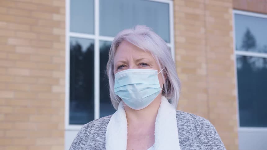 Elderly woman with grey hair and a light blue mask faces the camera with eye contact