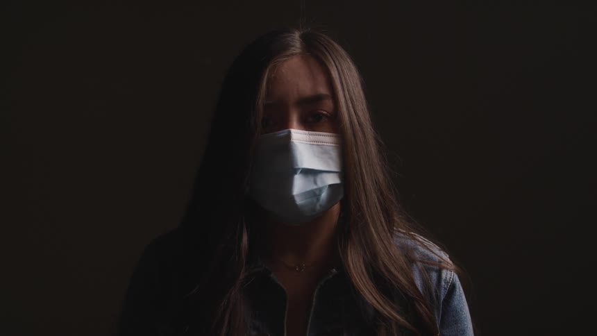 A young woman in a medical mask raises her head to look into the camera