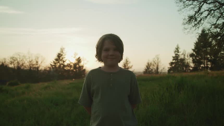 Boy with long hair and a grey t shirt stands on green grass and faces the camera with eye contact at dusk