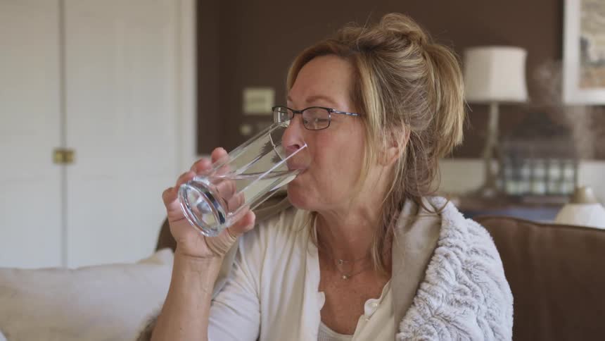 Middle aged woman with dirty blonde hair and glasses sits on a brown sofa and drinks a glass of water in the living room