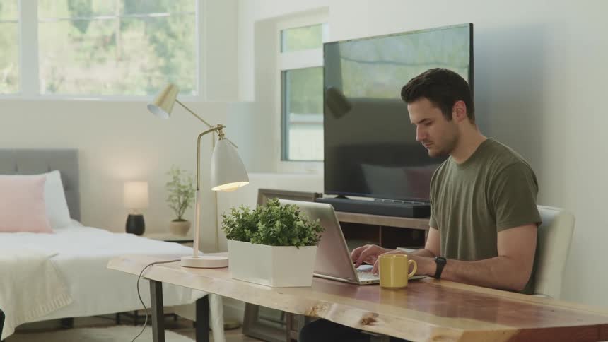 Man with a green t shirt sits at a wood desk uses a laptop in bedroom