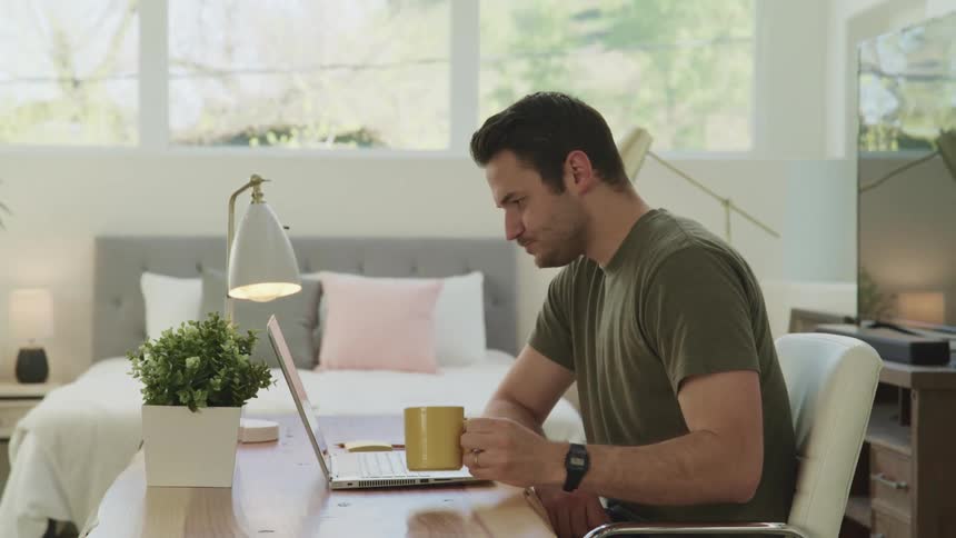 A man sitting at a desk in his bedroom talks with someone on his laptop as he drinks coffee