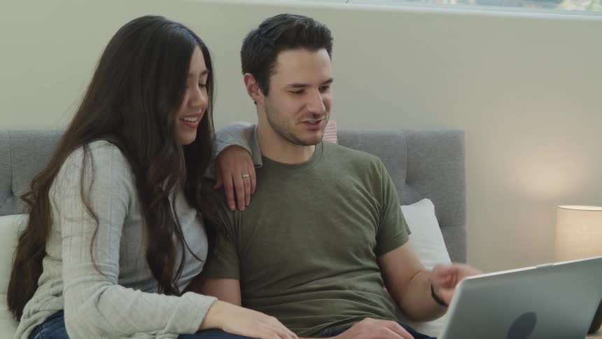 Young couple sits in the bed on a bedroom talking on a video call in a laptop