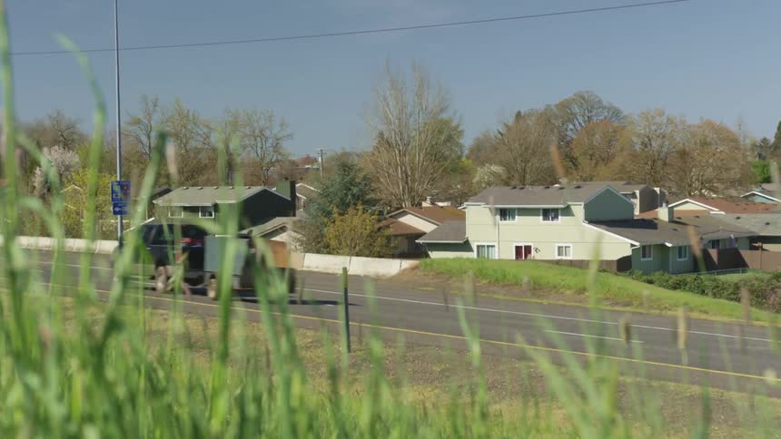 Through tall grass a pickup truck is shown pulling a trailer drives down a highway past a neighborhood