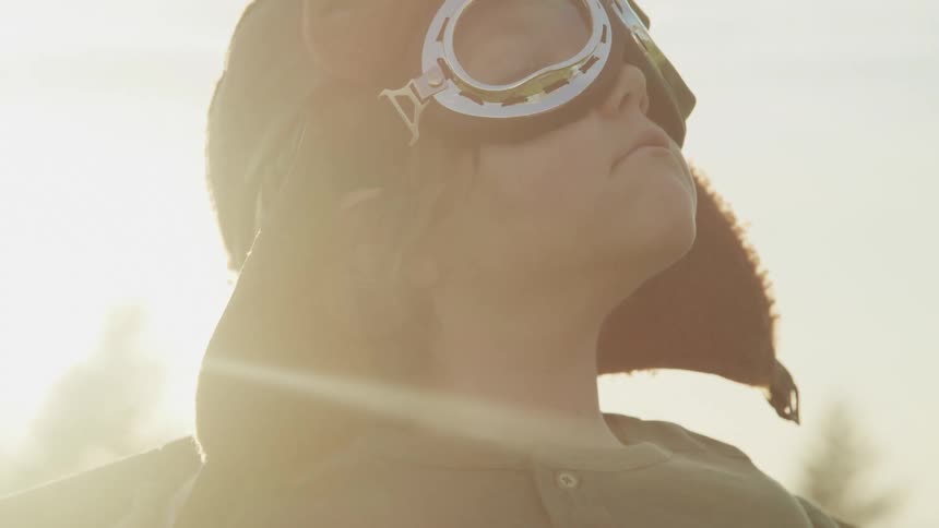 A child wearing aviation goggles and hat gazes at the sky as the sun shines behind them