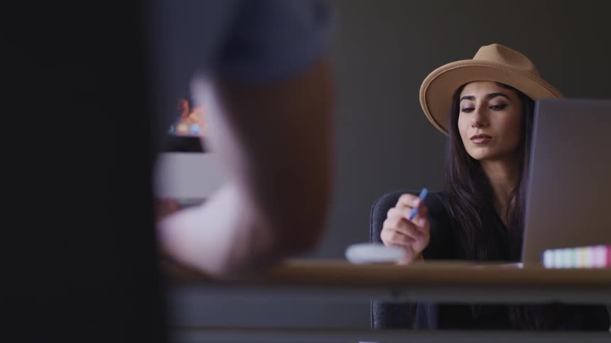 A woman sits in front of a laptop at a table while lost in thought for a moment before beginning to write on a piece of paper with a blue pen