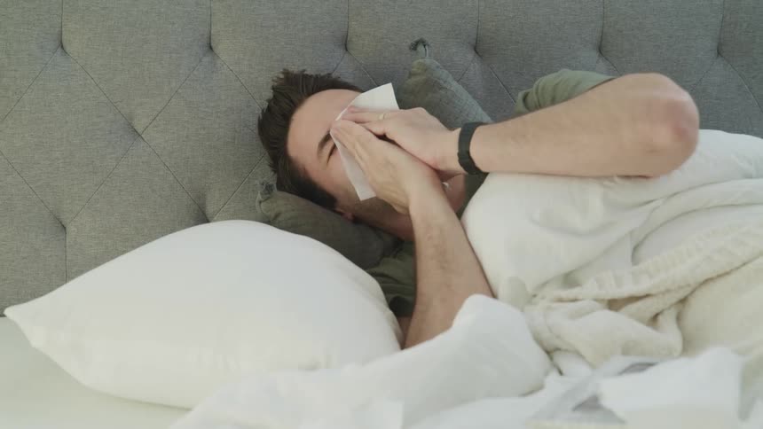 A sick man blows his nose while lying in bed