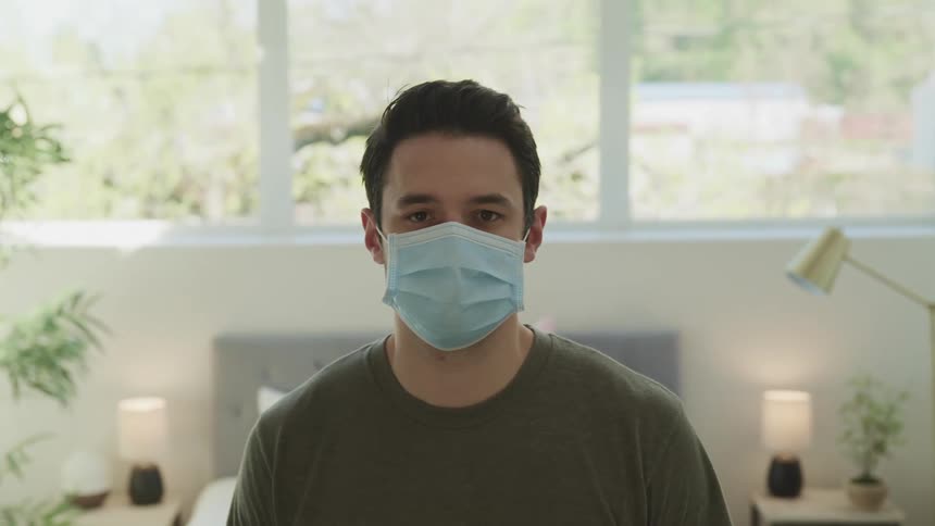 Man with a green t shirt and a light blue mask raises his head and faces the camera with eye contact in the bedroom