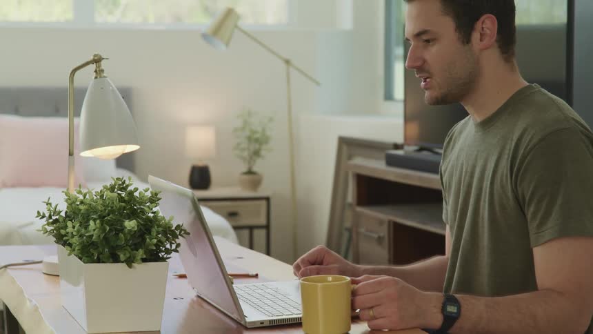 A man sitting at a desk in his bedroom talks with someone on his laptop as he hold a cup of coffee