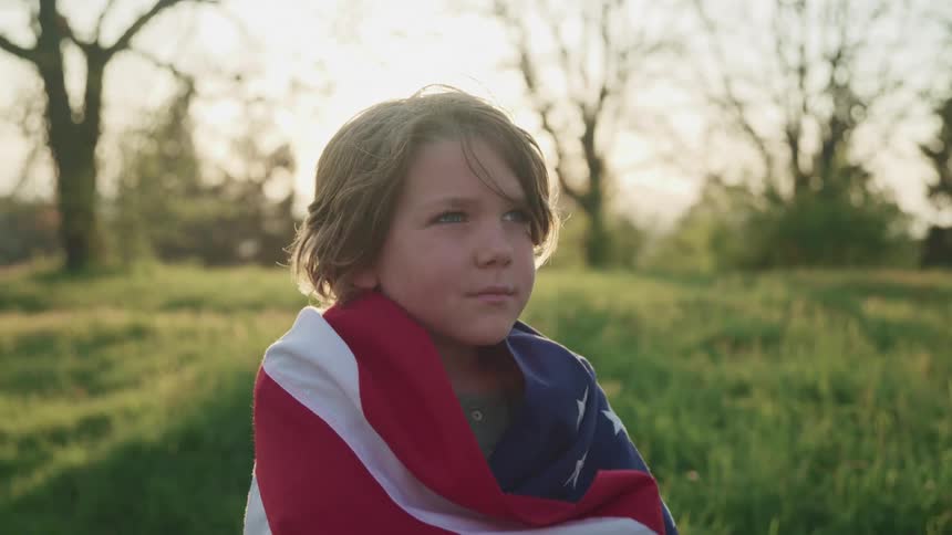 A young boy wrapped in an american flag stands in a field while gazing up at the sky