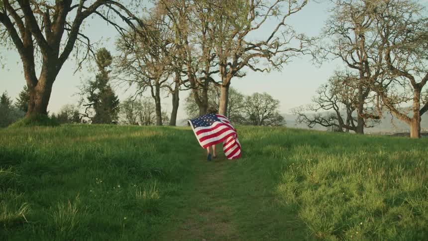 A child runs through a field with a fluttering american flag around his shoulders