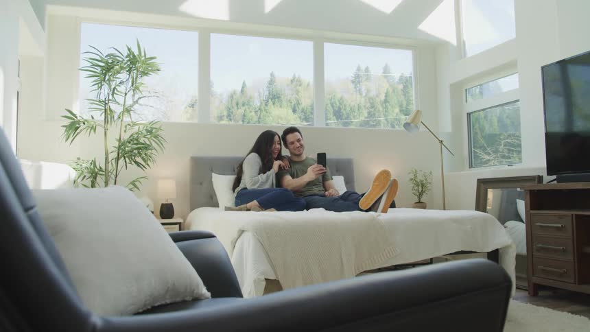 A woman and a man wave at the camera of a smartphone while they sit on a bed