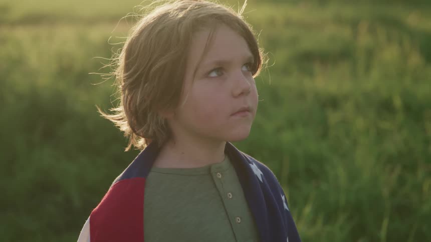 A boy stands outdoors in a field with an american flag draped over his shoulders