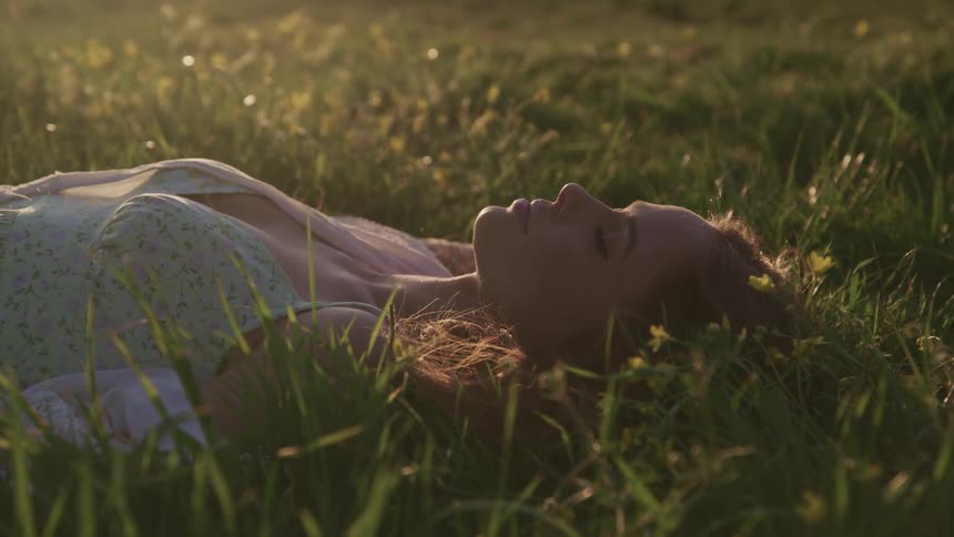Woman with brown hair lies down on green grass closes her eyes relaxing at sunset