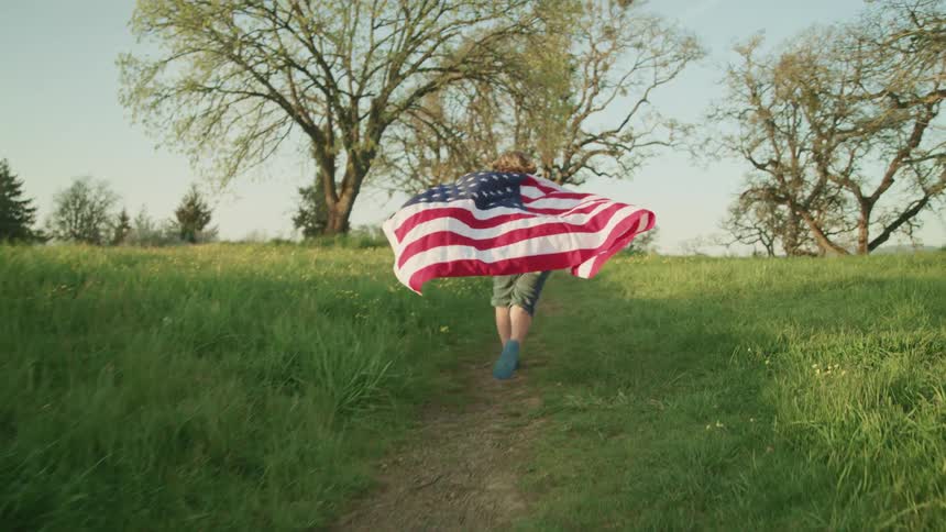 A person runs a pathway of a large open rural area with an american flag draped around their back